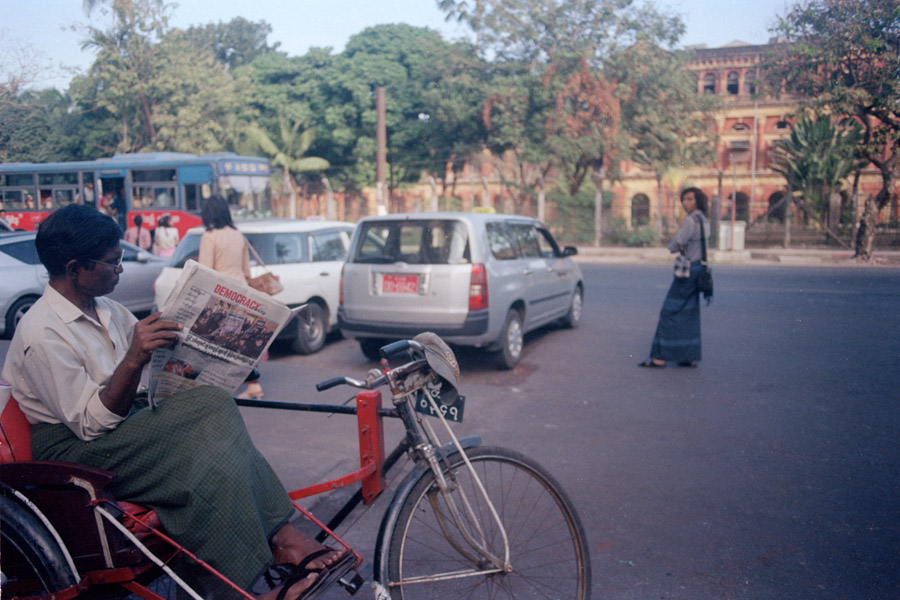 Yangon 2015: Traditional longyis on the men, beautiful colonial buildings, and trishaws for transportation, juxtaposed with the trishaw driver openly reading of a newspaper entitled 'Democracy Today.' Unthinkable just a few years ago under military rule. Despite this peaceful image, journalistic freedom is still in serious jeopardy in Myanmar.